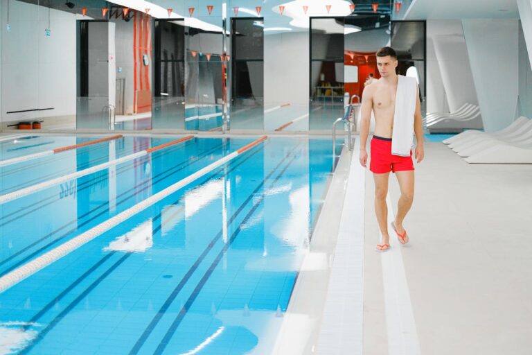 Shirtless young man walking poolside in an indoor swimming facility with a towel over his shoulder.